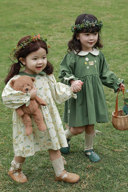 Girls in green and white dresses standing on grass, one holding a teddy bear and the other a basket.