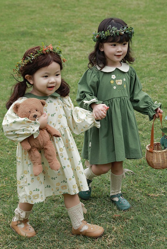 Girls in green and white dresses standing on grass, one holding a teddy bear and the other a basket.